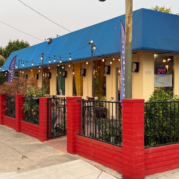 Restaurant exterior with bright blue awning, red brick patio walls, string lights, outdoor seating, and an illuminated open sign.